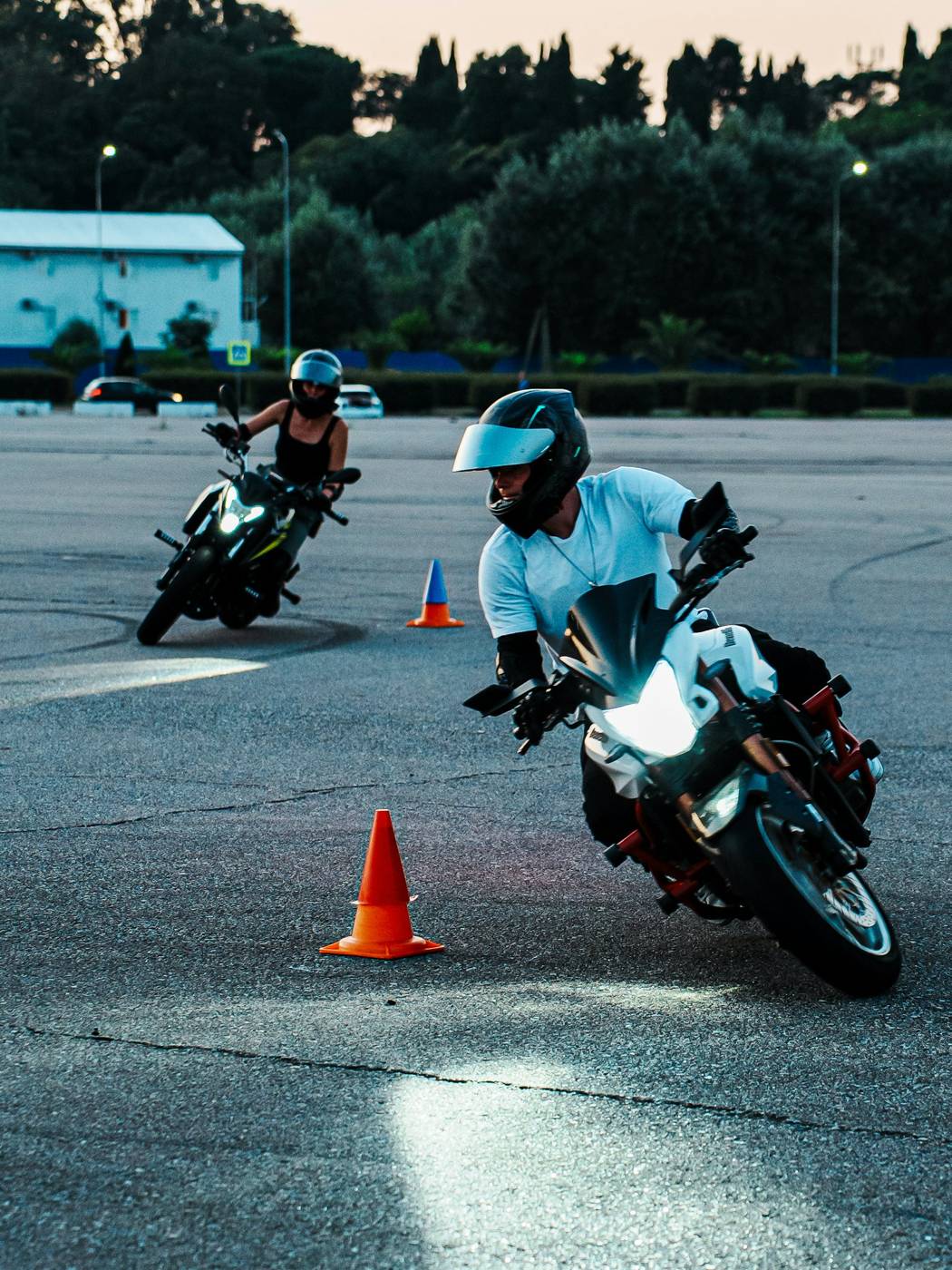 Beginner motorcycle riders practicing slow turns around orange cones on a closed practice lot